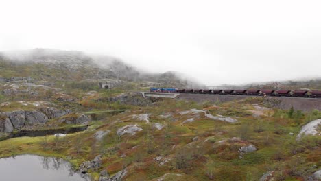 aéreo: tren de mineral en la estación de søsterbekk, cerca de la frontera entre suecia y noruega en el norte de laplandia cruzando un puente en un lago y entrando en un túnel en un paisaje brumoso