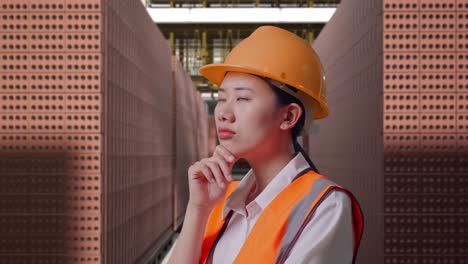 close up side view of asian female engineer with safety helmet thinking about something and looking around while standing with red brick packed in stacks are stored