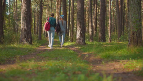 back view of two sisters walking along forest trail surrounded by lush greenery, one wears blue hair tie, carrying black backpack, while other has red backpack, tall trees line path