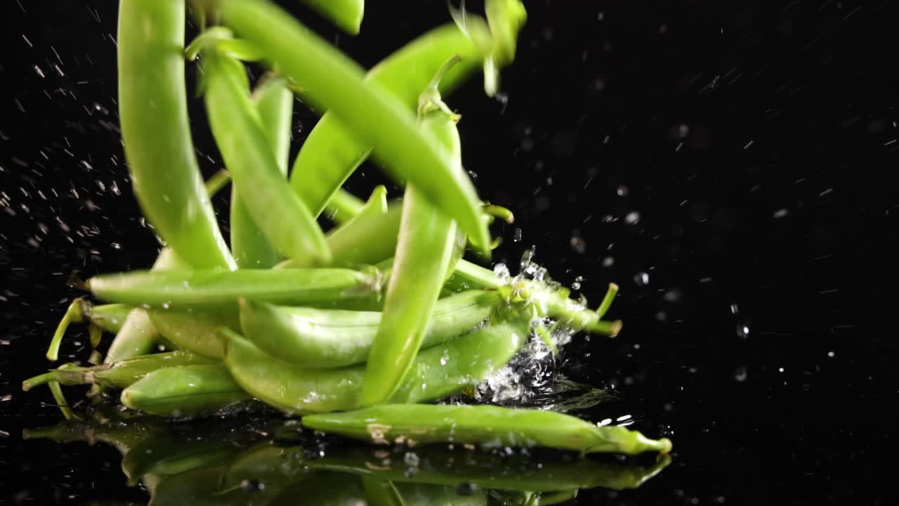 Green peas fall onto a reflective black surface, creating dynamic splashes and movement, captured in slow motion