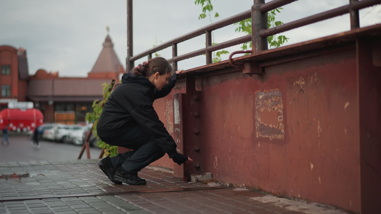 Junge Frau auf nasser Metallbrücke, hält inne und inspiziert den Gehweg, dann hockt sie sich hin, um Schnürsenkel zu binden, verrostetes Geländer und Marktarchitektur im Hintergrund, Regentropfen und strukturierte Oberfläche, authentisches Detail