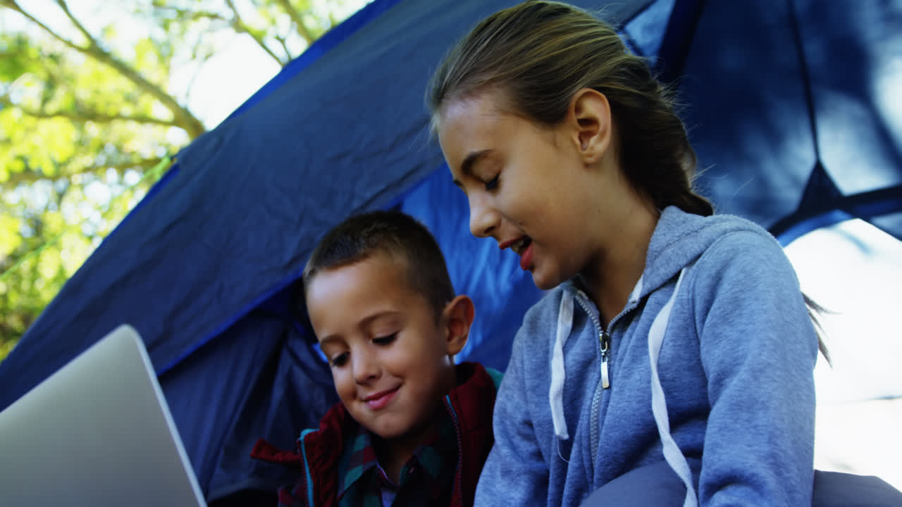 hermanos usando computadora portátil fuera de la tienda en el campamento