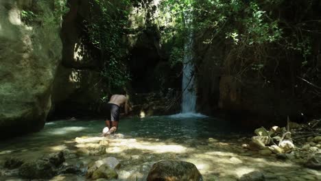 joven caucásico sin camisa y descalzo con traje de baño negro camina hacia la piscina de montaña fresca junto a una cascada y se sumerge, antena baja hacia atrás