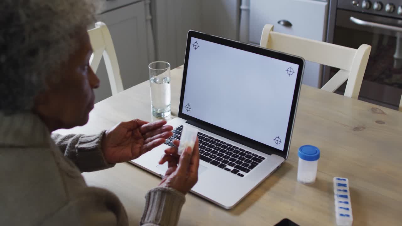 Senior african american woman holding medication container having a videocall on laptop at home