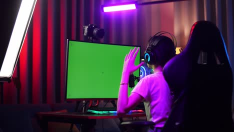 back view of asian girl streamer feeling disappointed fails game over on green screen computer. live stream on desk illuminated by rgb led strip light