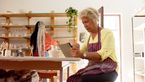 Happy-biracial-female-potter-with-gray-hair-using-tablet-in-pottery-studio,-slow-motion