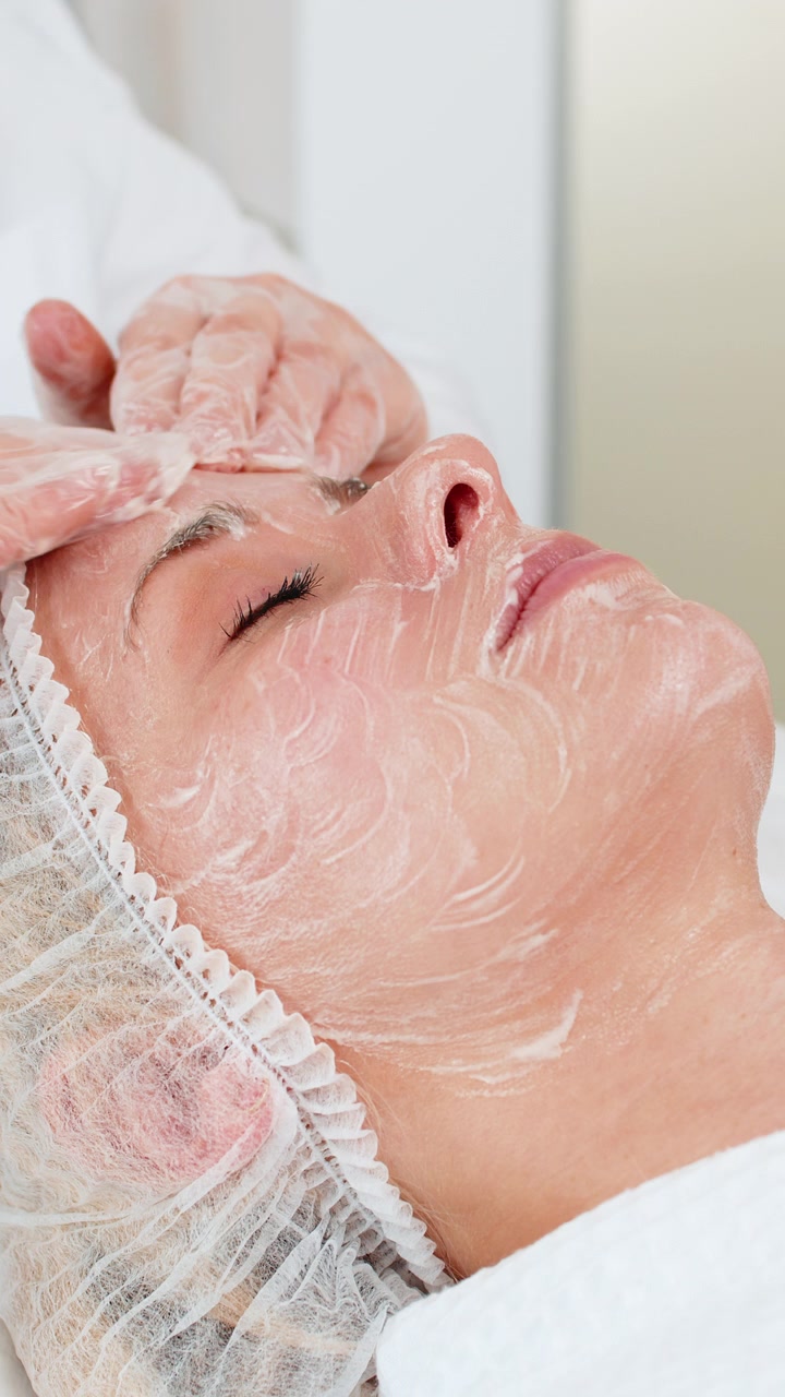 Cosmetology clinic facial peeling beautician rubbing moisturizing cream on middle aged woman skin