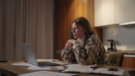 a pensive woman in glasses looks at the laptop screen and concentrates and comes up with a development strategy. schedules and plans on paper are on the table