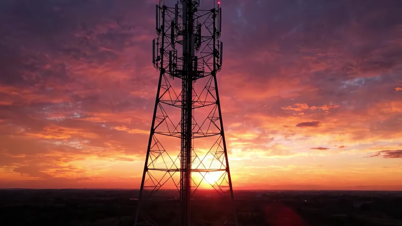 Un tramonto mozzafiato sagoma una torre delle comunicazioni contro uno sfondo di nuvole arancioni, rosa e viola vibranti, racchiudendo la bellezza della natura e della tecnologia.