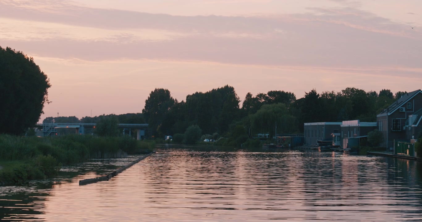 Безмятежный водный путь на закате, отражающий нежные розовые и лавандовые оттенки, с деревьями и небольшими строениями вдоль берегов