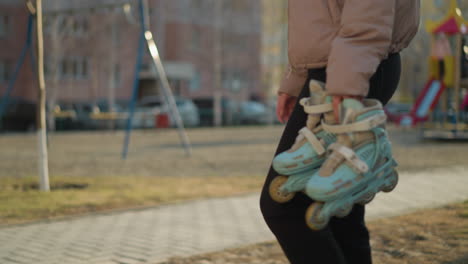 a faceless person is walking through a sunlit park, holding a pair of rollerblades in one hand. the individual is dressed in a peach jacket and black trousers, with blurred houses in the background