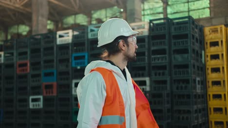 A-brunette-man-with-a-beard-in-a-protective-suit-goggles-and-a-helmet-in-an-orange-vest-walks-along-black-and-yellow-boxes-for-sorting-and-recycling-waste-at-a-large-factory