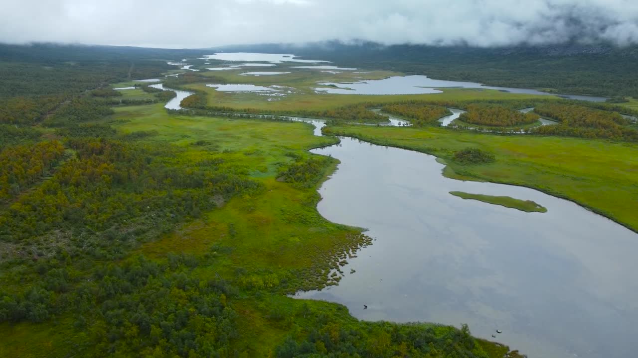 Aerial drone footage flying slowly over Sweden wetlands or marshlands during a cloudy day with large lakes and rivers visible in the foreground with green moss and forests surrounding the water.