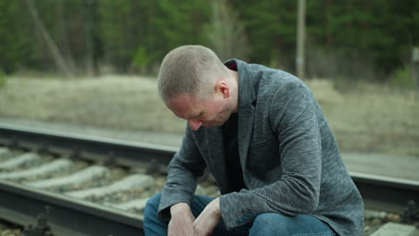 a close view of a man in a gray jacket, blue jeans, and canvas shoes, handling a handgun while sitting beside a railway track, the background shows a blurred view of a green forest