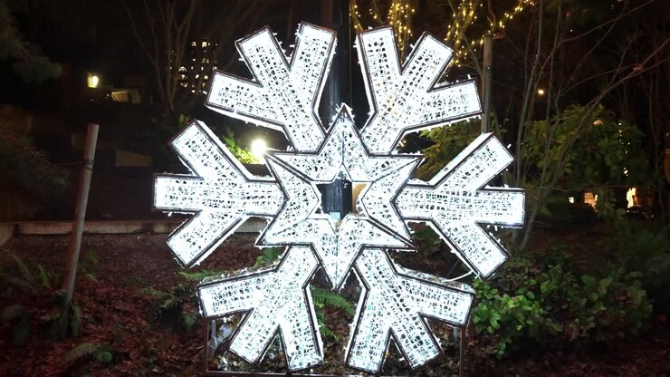 Centred push in on large white snowflake christmas decoration sat on a verge in a local city park in the Canadian city of Vancouver at night causing a festive glow