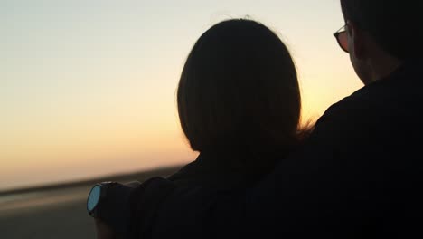 pareja viendo la puesta de sol y abrazándose en la playa, de mano cerca