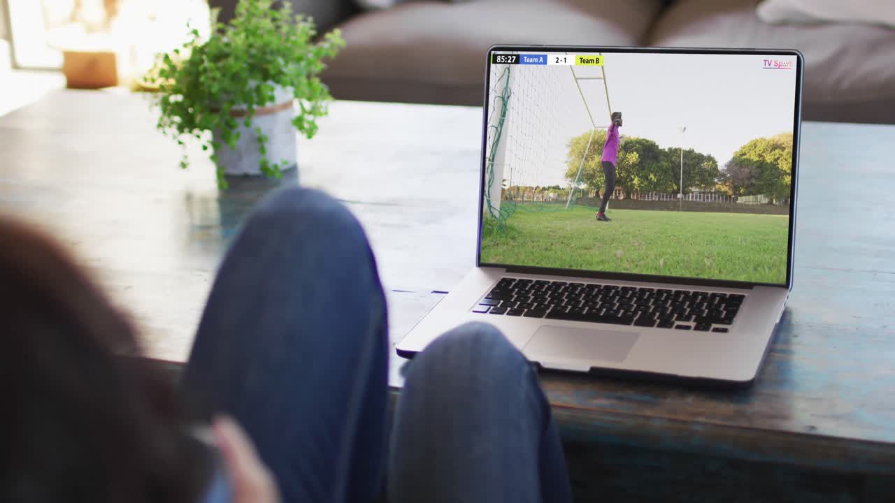 Man using laptop with african american male soccer player playing match on screen