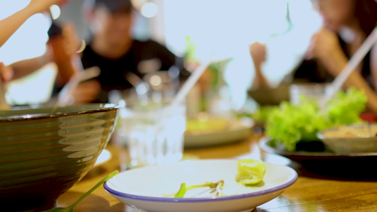 A lively group enjoys Vietnamese cuisine at a bustling table in Bangkok, with dynamic interactions and vibrant lighting