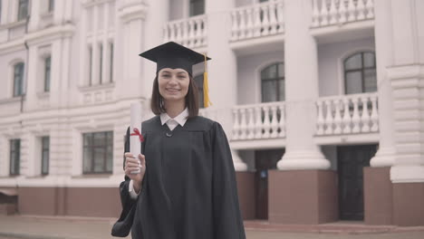 linda estudiante de posgrado en bata y gorra con diploma mirando a la cámara y celebrando