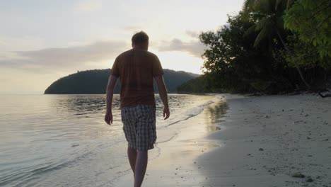 A-man-strolls-along-the-sandy-beach-of-Kri-Island-in-Raja-Ampat,-Indonesia,-at-sunset