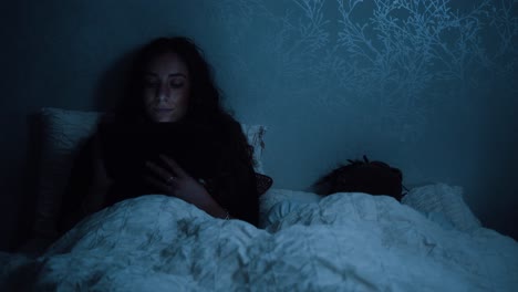 young woman sits up in bed at night while watching a tablet device