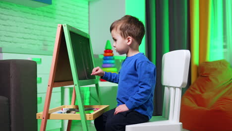 lovely toddler kid sits at the blackboard at home. little kid draws with a chalk excitedly.