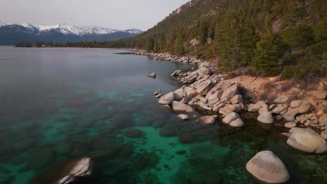 sunny shoreline mid winter day sand harbor secret beach south lake tahoe aerial drone california large boulder rocks crystal clear glassy turquoise fresh water snowy northstar ski resort forward up