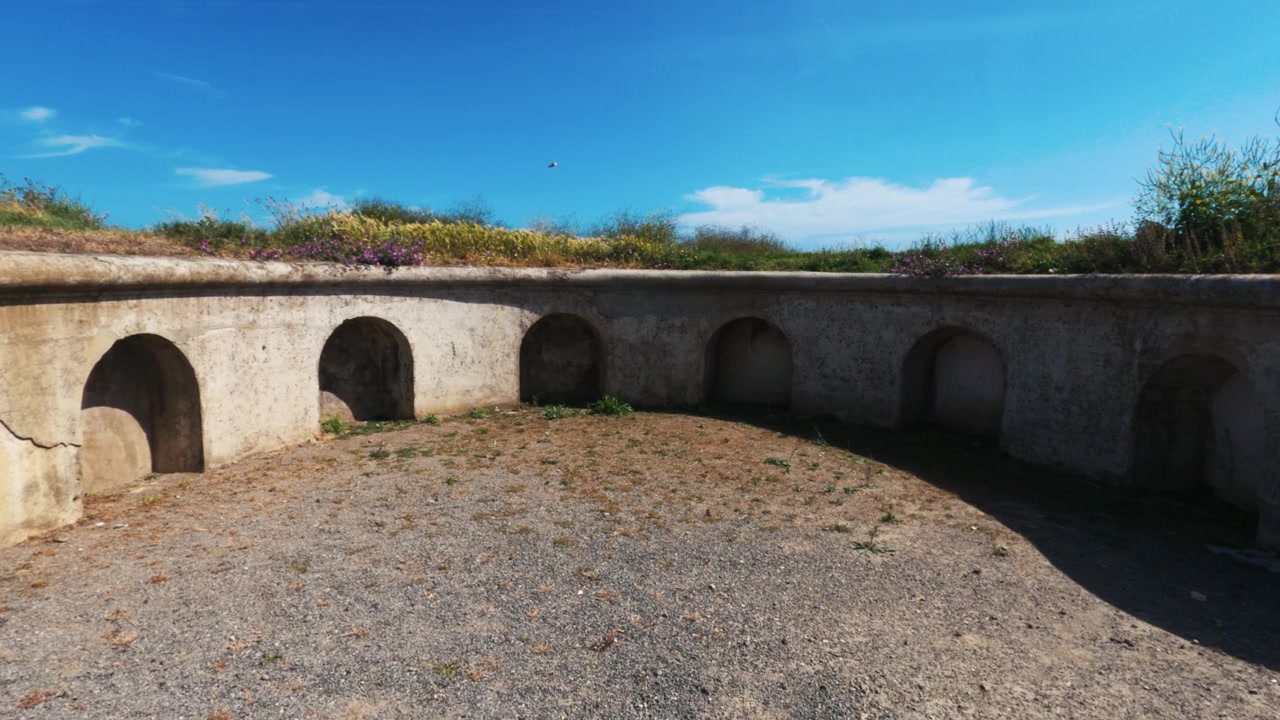 Imagem estática ao nível do solo de um muro de pedra curvo com nichos em arco numa fortificação de artilharia histórica no Cerro Santa Catalina, Gijón.