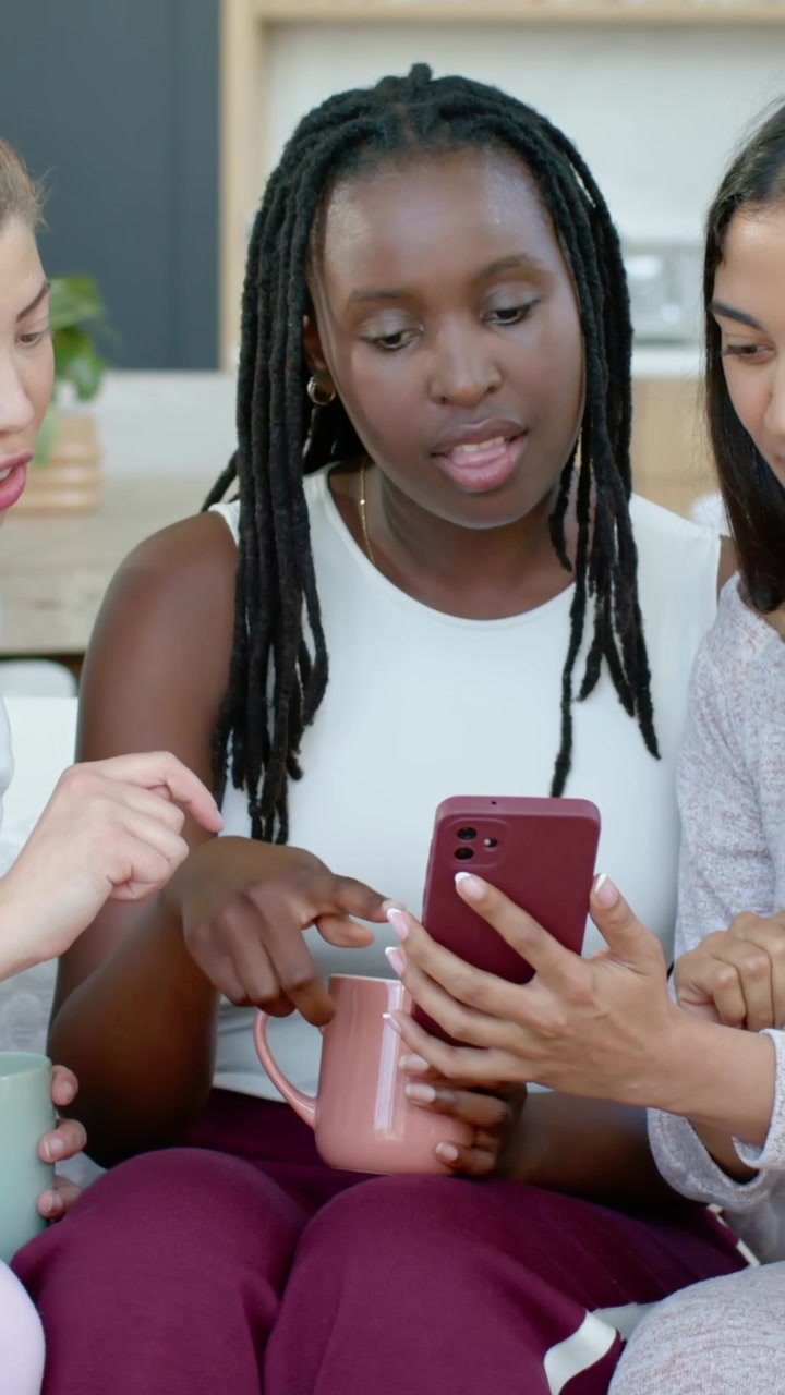 Vertical video: Right woman showing red phone, trio viewing screen on couch, center holding mug