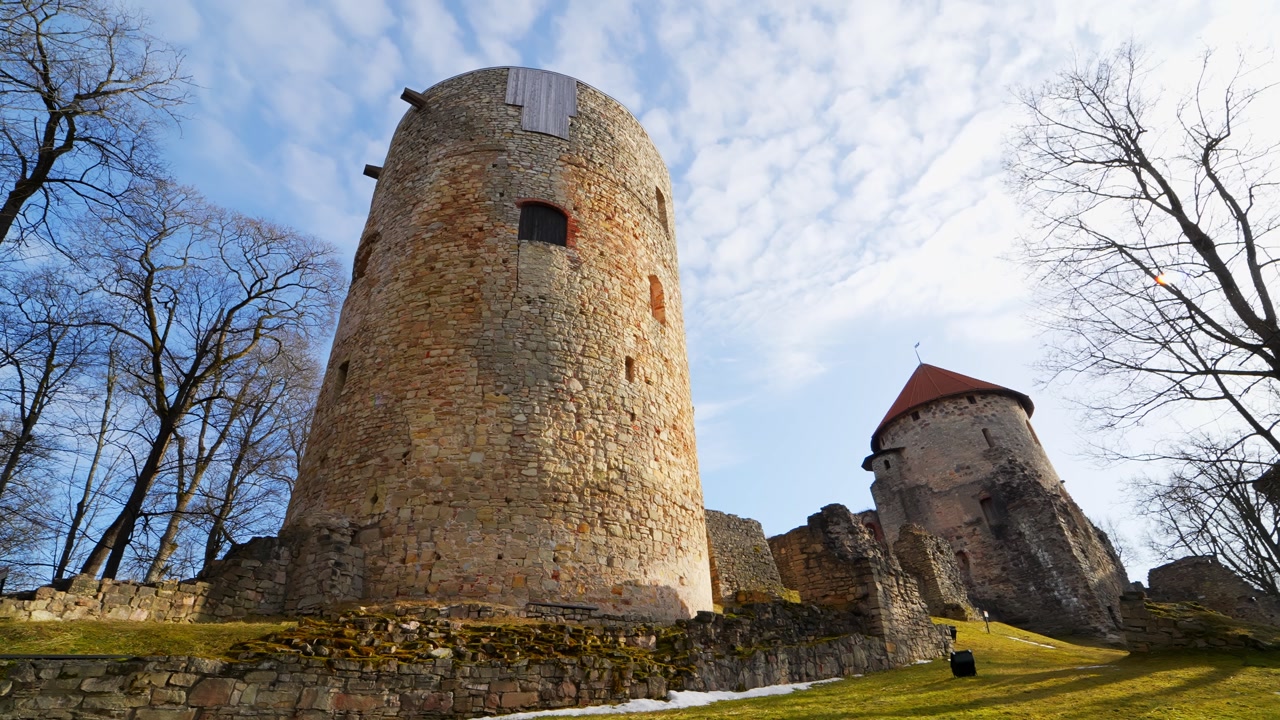 Low-angle view of medieval castle towers and stone ruins in bright spring light, with bare trees, blue sky, and historic fortress architecture on a grassy slope.