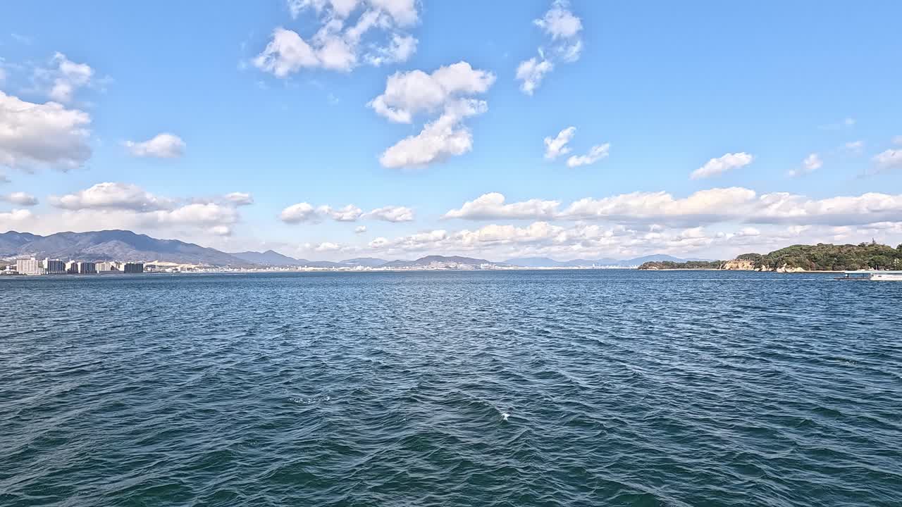 Serene ocean view from the deck of a ferry crossing Hiroshima Bay toward Miyajima Island in Japan, with a mountainous coastline in the distance under a partly cloudy sky.