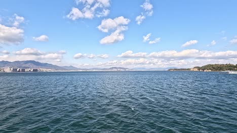 serene ocean view from the deck of a ferry crossing hiroshima bay toward miyajima island in japan, with a mountainous coastline in the distance under a partly cloudy sky.