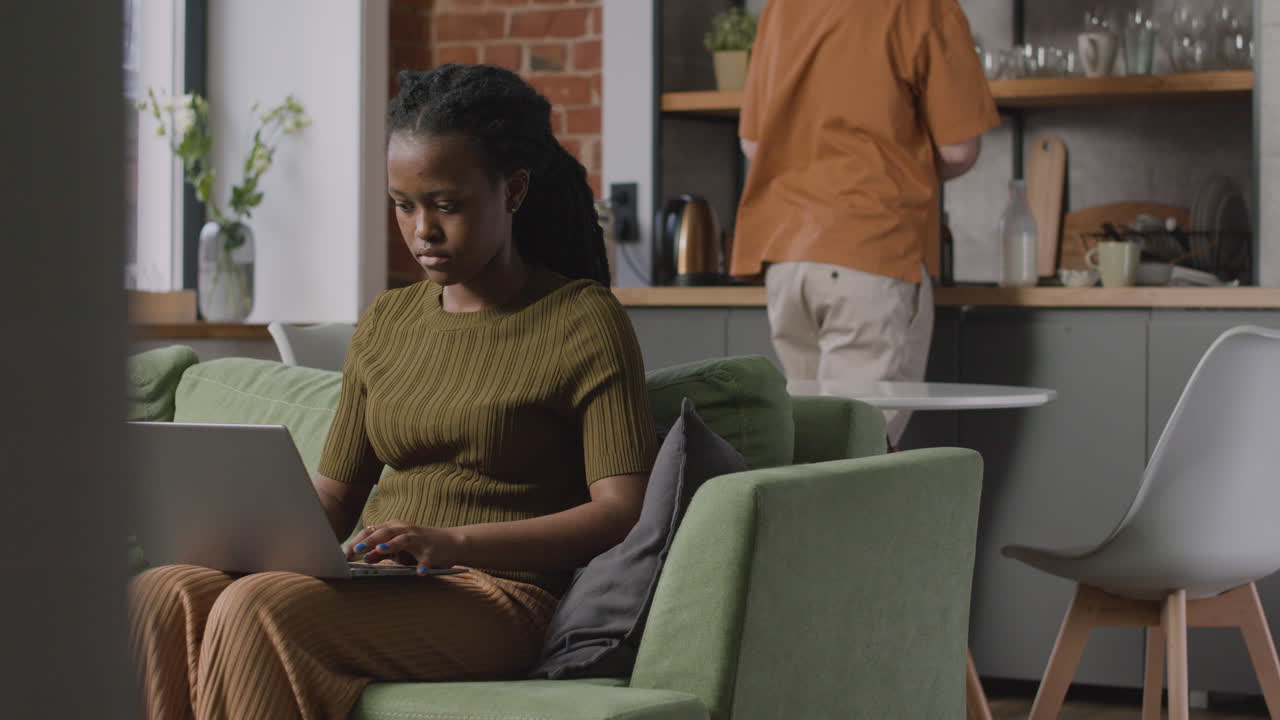 Girl Working On Laptop Computer Sitting On Sofa While Her Male Roommate Cleaning The Kitchen Table