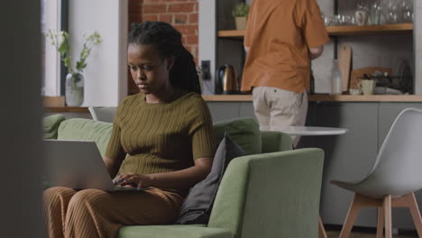 Chica-Trabajando-En-Una-Computadora-Portátil-Sentada-En-Un-Sofá-Mientras-Su-Compañero-De-Cuarto-Limpia-La-Mesa-De-La-Cocina