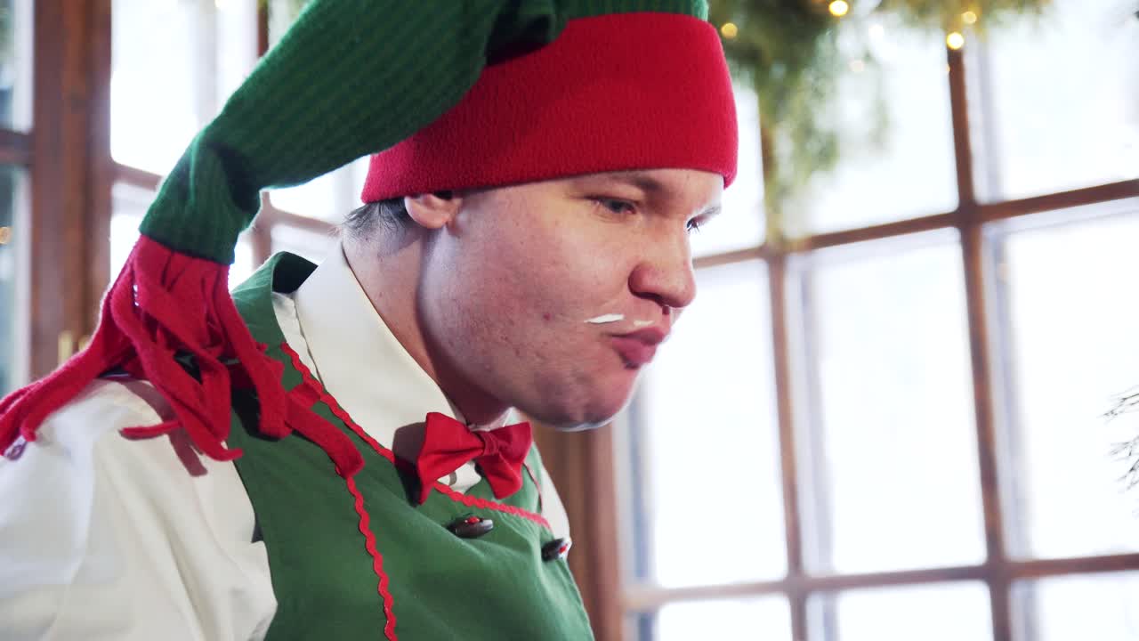 Funny elf with dirty face eats quickly. Hungry male elf in red and green hat eats and drinks indoors. Close-up