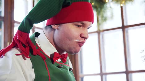 funny elf with dirty face eats quickly. hungry male elf in red and green hat eats and drinks indoors. close-up