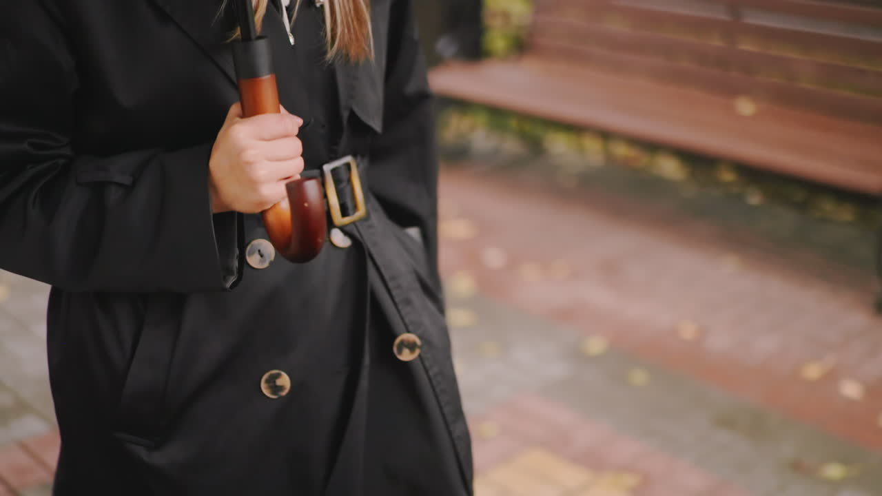 Close-up of woman hand holding wooden handle umbrella while wearing black coat on rainy autumn day, blurred pavement and fallen leaves in background, detail shot of stylish urban lifestyle
