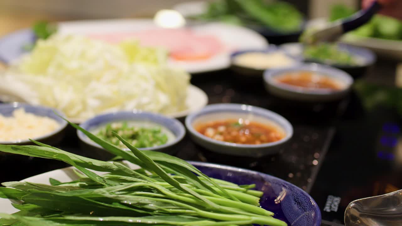 Hand selects dipping sauce among fresh vegetables, sliced meat, and condiments for hotpot meal