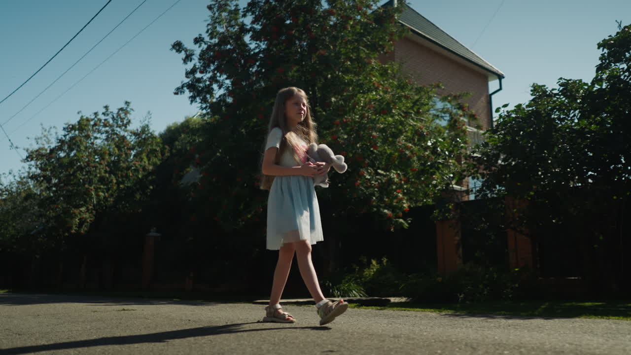 Young girl walks outdoors along quiet residential street on sunny day, holding plush bunny in hand. She is dressed in soft blue dress, surrounded by trees and houses casting long shadows