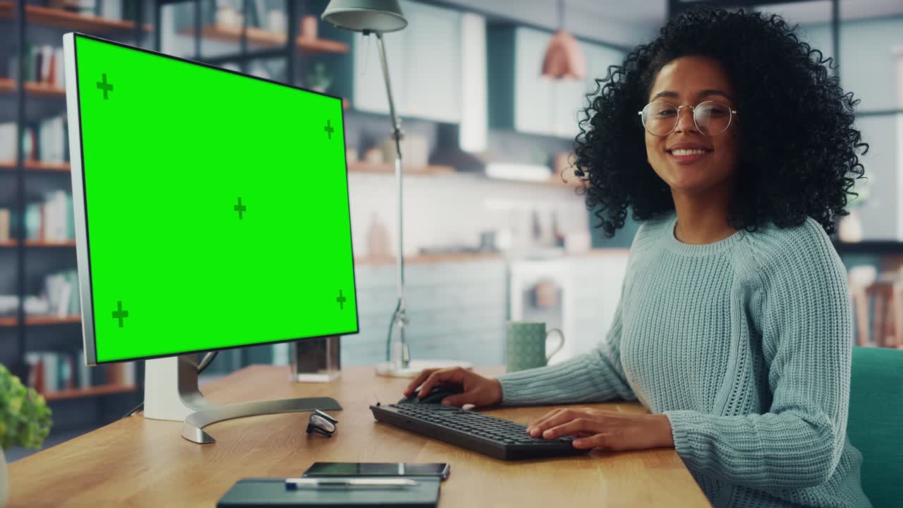 Latina Female Specialist Working on Desktop Computer with Green Screen Mock Up Display at Home Living Room while Sitting at a Table. Happy Freelancer Female Looks at the Camera and Smiles.