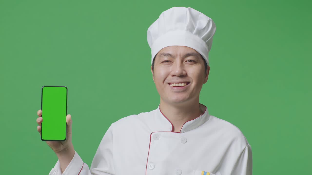 녹색 스크린 배경 스튜디오에 서 있는 동안 카메라에 녹색 화면 스마트폰을 보여주고 미소 짓는 아시아 남자 셰프의 클로즈업
