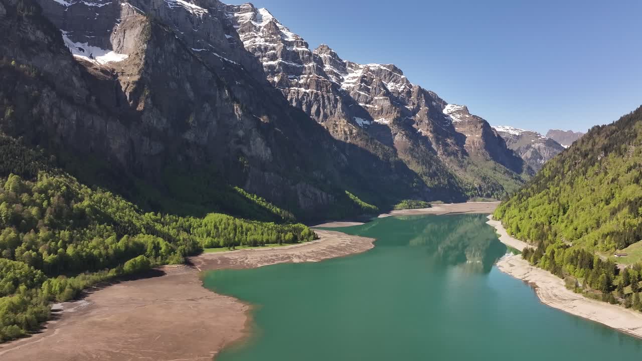 Aerial - turquoise Klöntalersee lake surrounded by green forests and snow-capped Swiss Alps