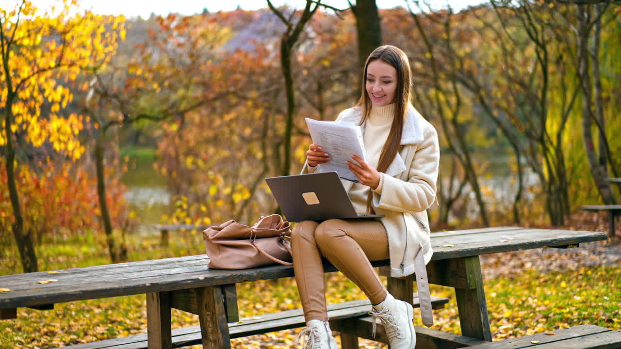 Smiling long-haired brunette lady looking through the papers in her hands. Woman sits on table in the park with laptop on her knees and having some remote work. Autmn nature at backdrop.