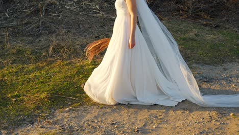 elegant slow-motion close-up of a bride walking at sunset, holding dry grass—head not in frame.
