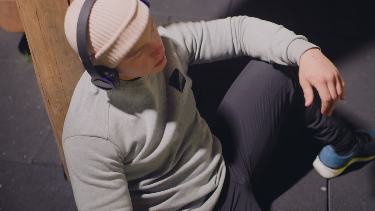 man som vilar på gymgolvet, basketboll, hörlurar och mössa, lutad mot en betongpelare under dämpad belysning, sporttröja och joggingbyxor, gympaskor på gummimatta, andas efter träningspasset
