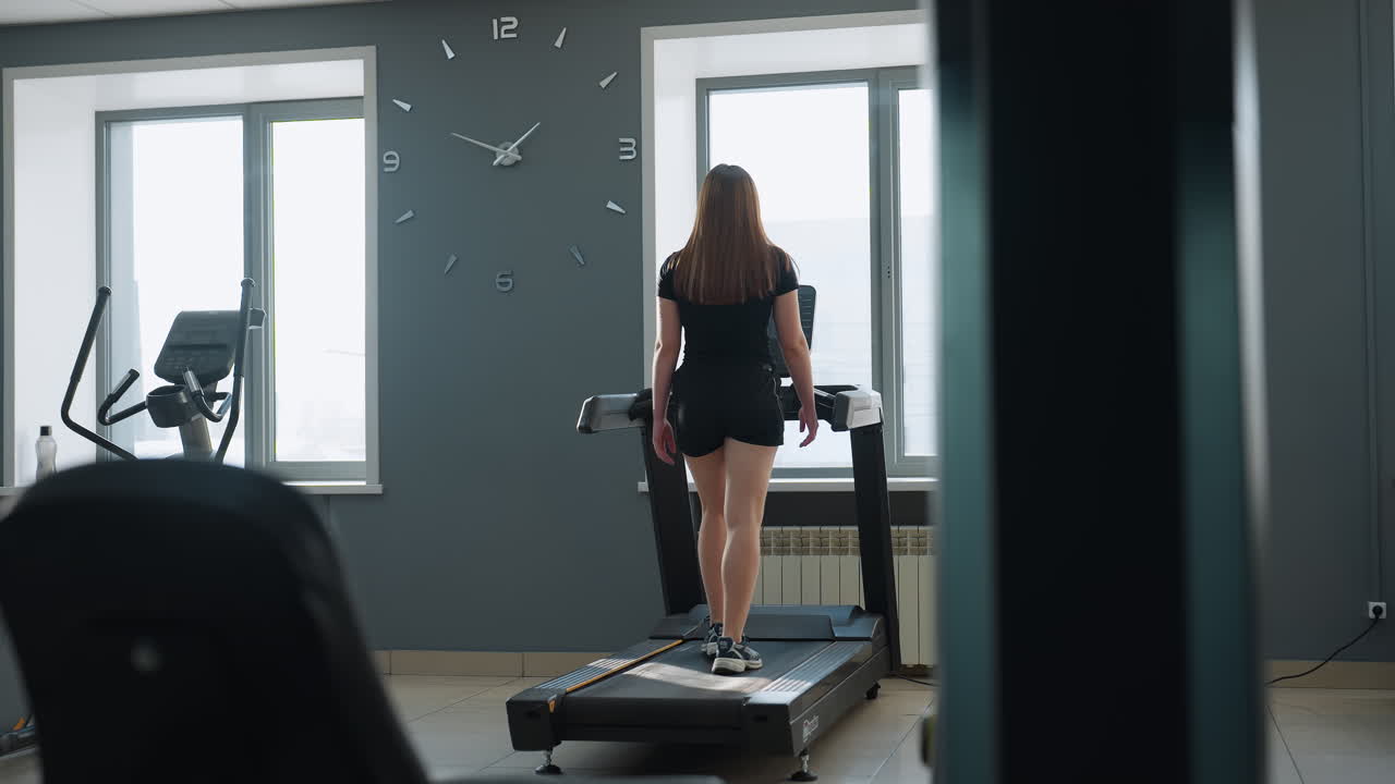 rear view of woman exercising on treadmill in modern fitness studio with sunlight through windows, elliptical machine and water bottle visible in background, treadmill plugged into socket