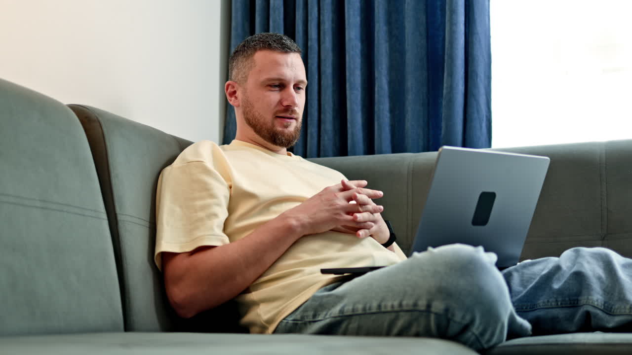 Man sits on sofa and uses laptop while focusing on tasks at home during late afternoon hours in a comfortable living room setting