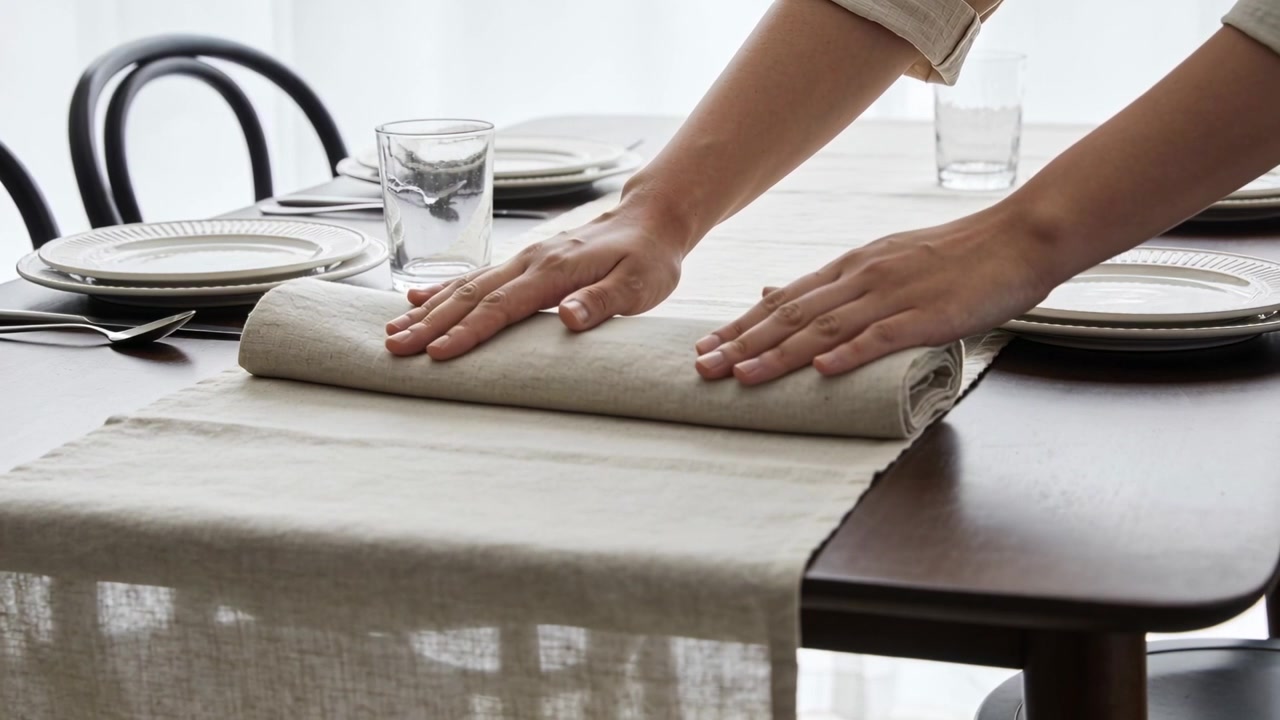 Mãos com as mangas arregaçadas desenrolando e ajeitando um caminho de mesa de linho sobre a mesa de jantar, preparando para a refeição.