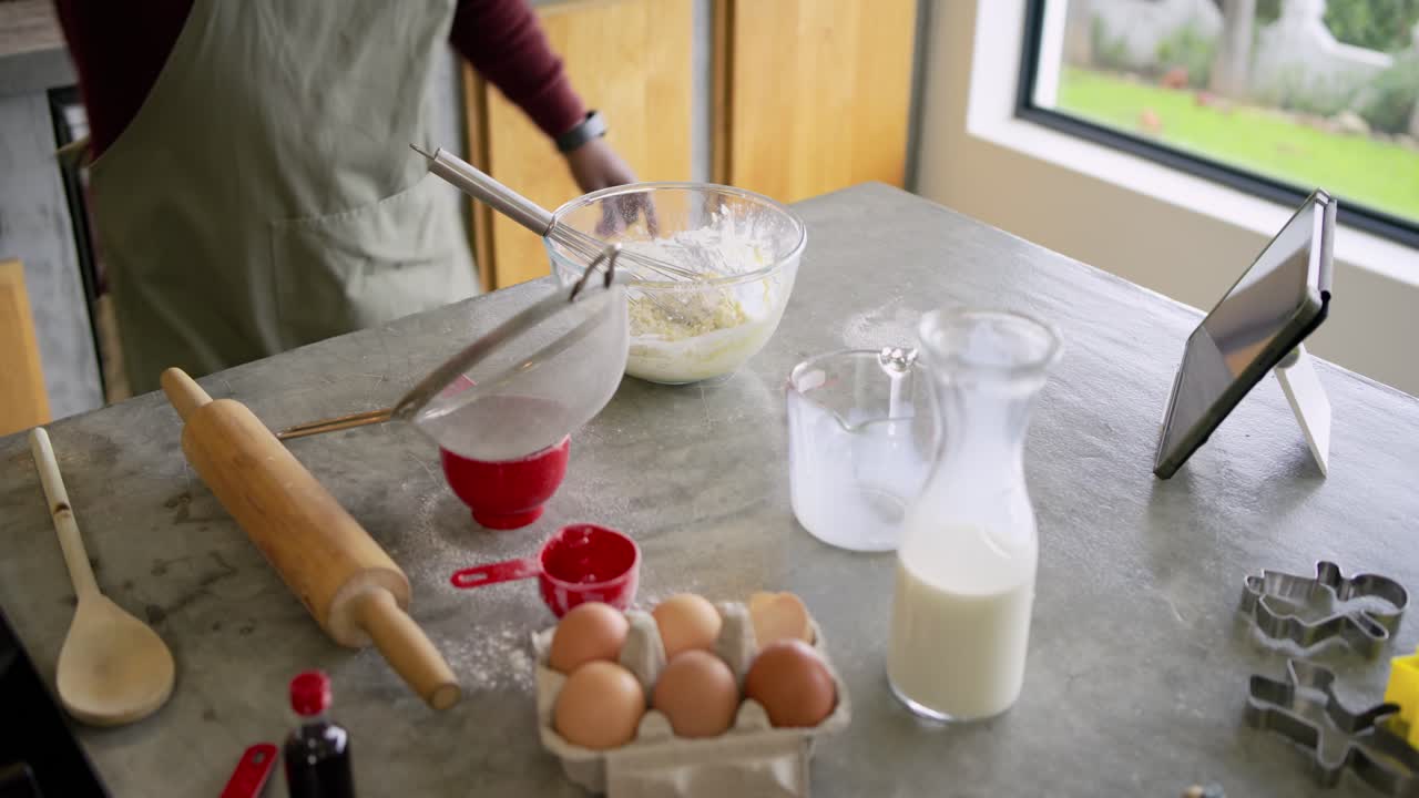 African American man wearing green apron and Santa hat checking recipe in kitchen, whisking batter