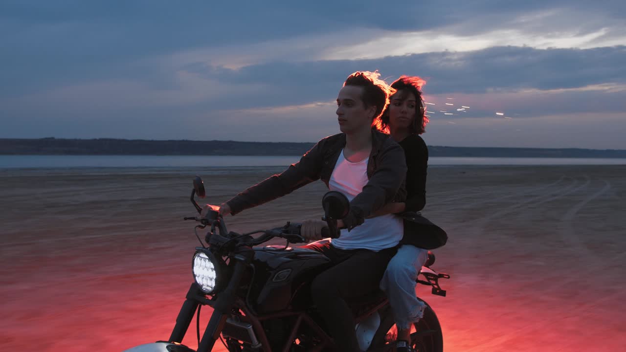 해변에서 일몰 후 빨간색 불타는 신호 불과 함께 빈티지 오토바이를 타고 있는 젊은 부부, 느린 동작